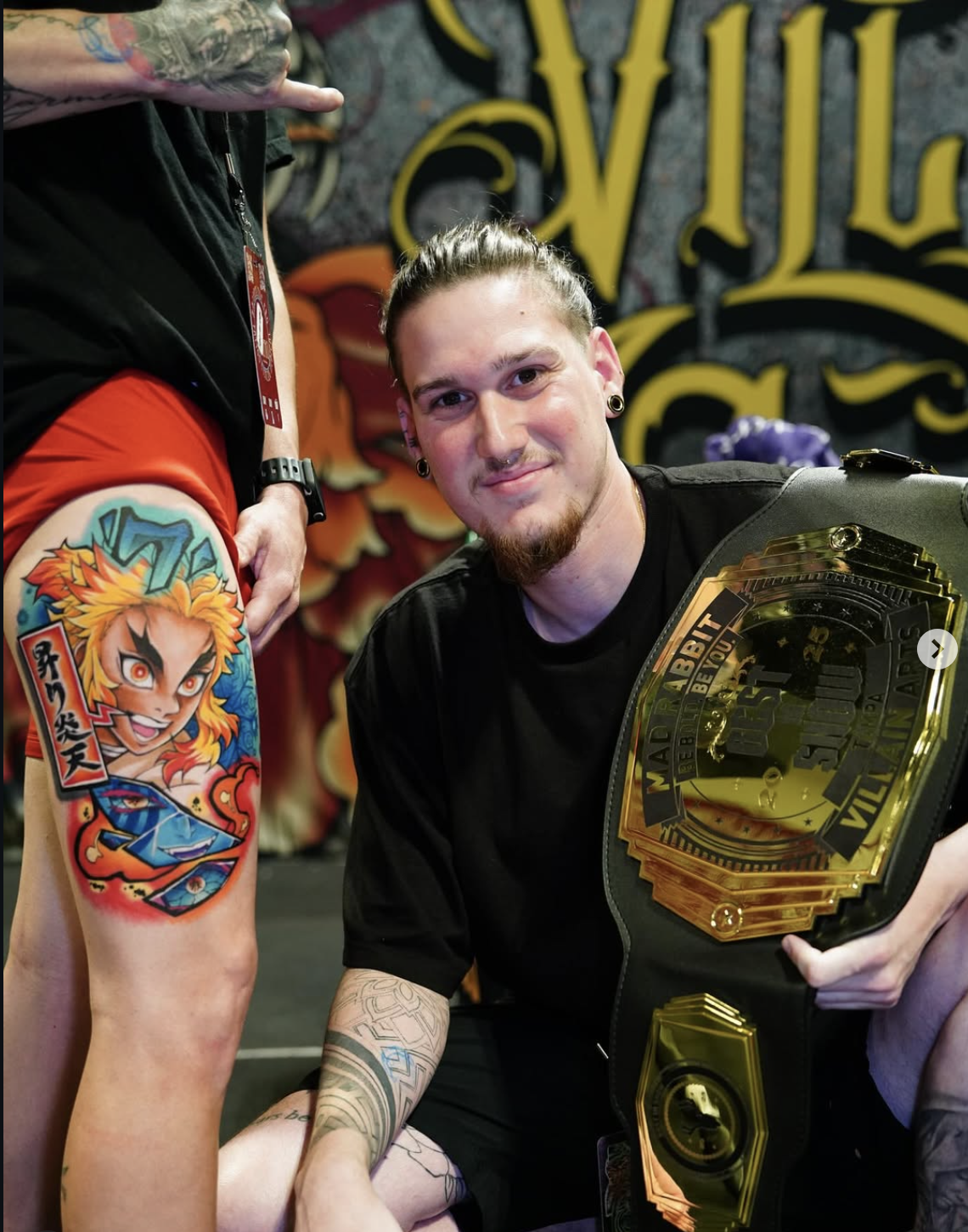 Osiel holding a tattoo convention championship belt beside a finished color tattoo